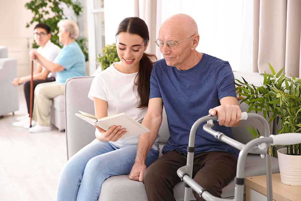 A Young Woman Holding a Book Sitting Beside An Elderly Man Holding On To A Walker What Do Hospice Volunteers Do